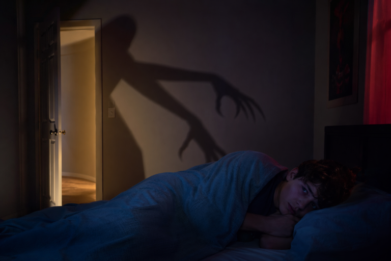 A Nightmare on Elm Street scene with a terrified teen in bed as a clawed shadow looms across the bedroom wall in eerie red and blue light.