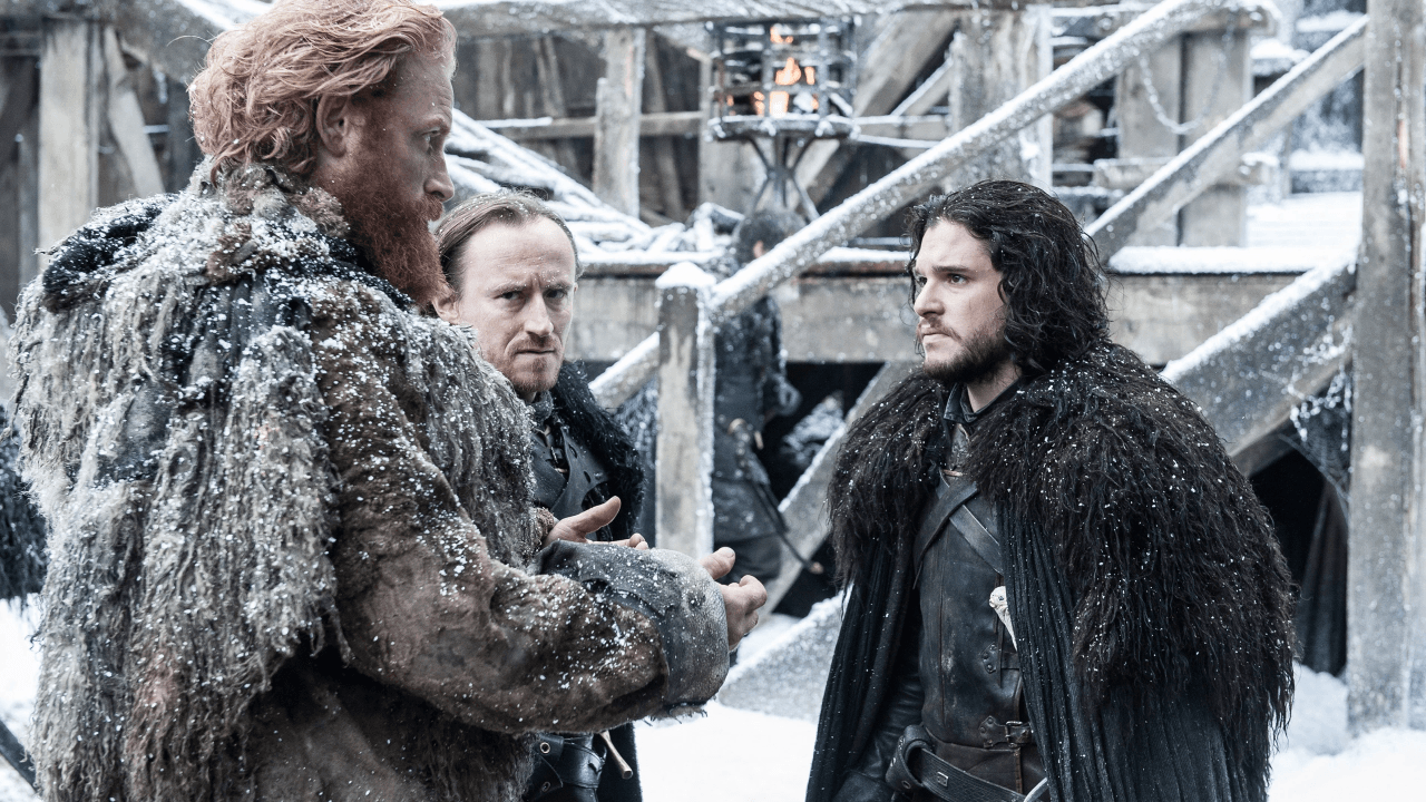 A commander in a black cloak stands before supplies and recruits at a snow-covered fortress wall in the Game of Thrones S5E7 "The Gift" episode guide scene.